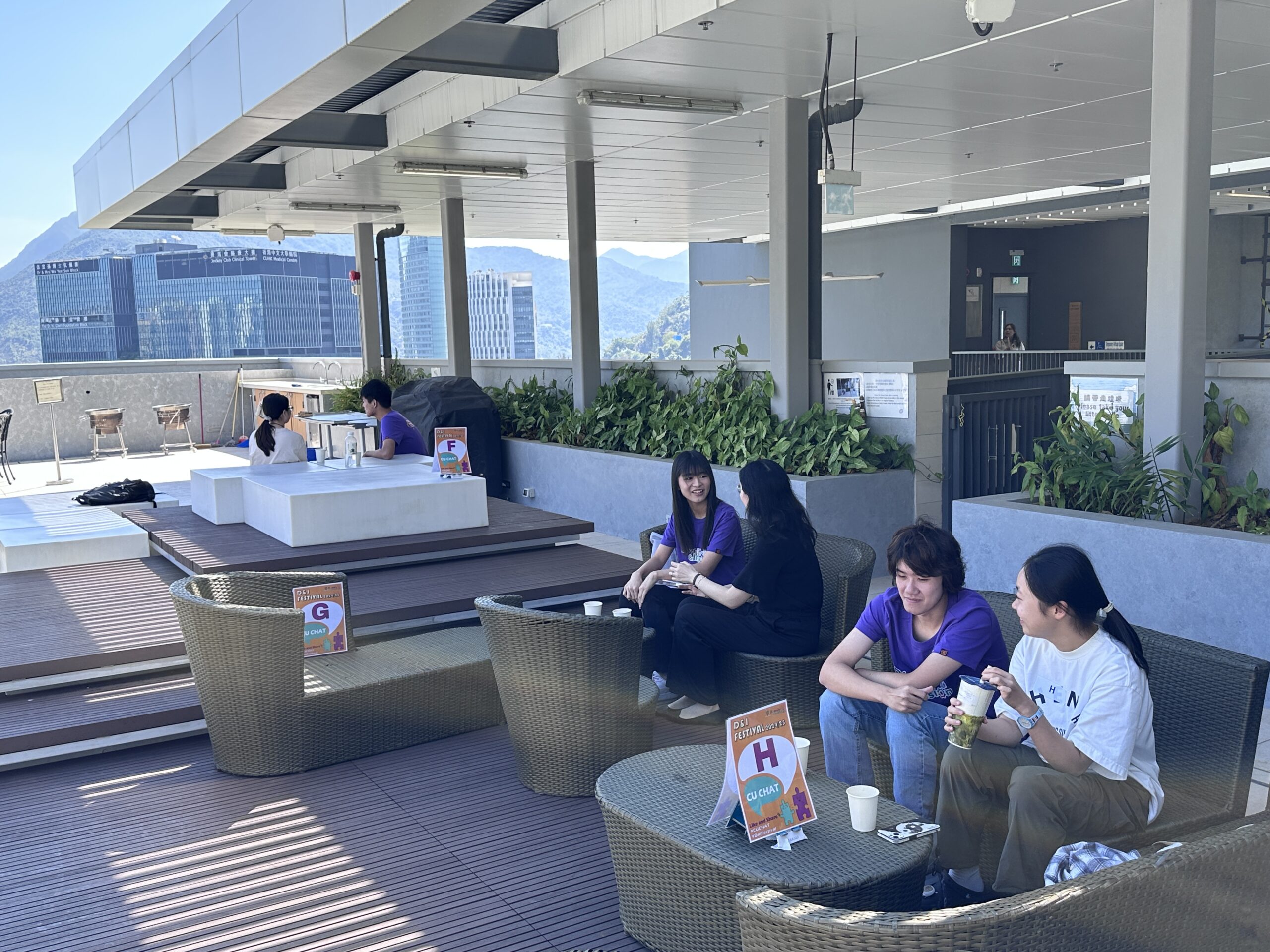 Ambassadors and participants enjoy a conversation while sitting on a balcony with scenic views of mountains and campus buildings. All of them are casually dressed and drinking beverages.