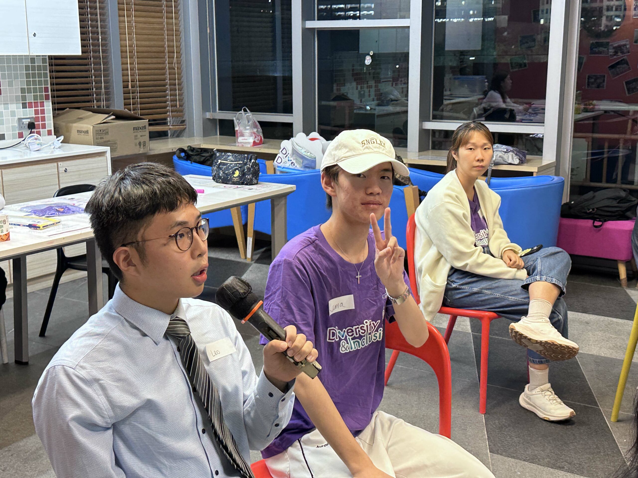 Three D&I ambassadors are seated in a room, engaged in discussion. One holds a microphone, while another makes a victory sign.