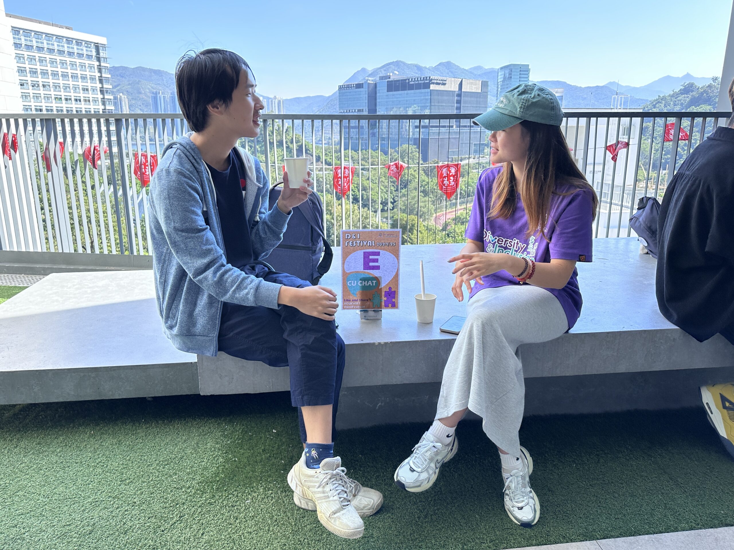 The ambassador and a participant enjoy a conversation while sitting on a balcony with scenic views of mountains and buildings. Both are drinking beverages.