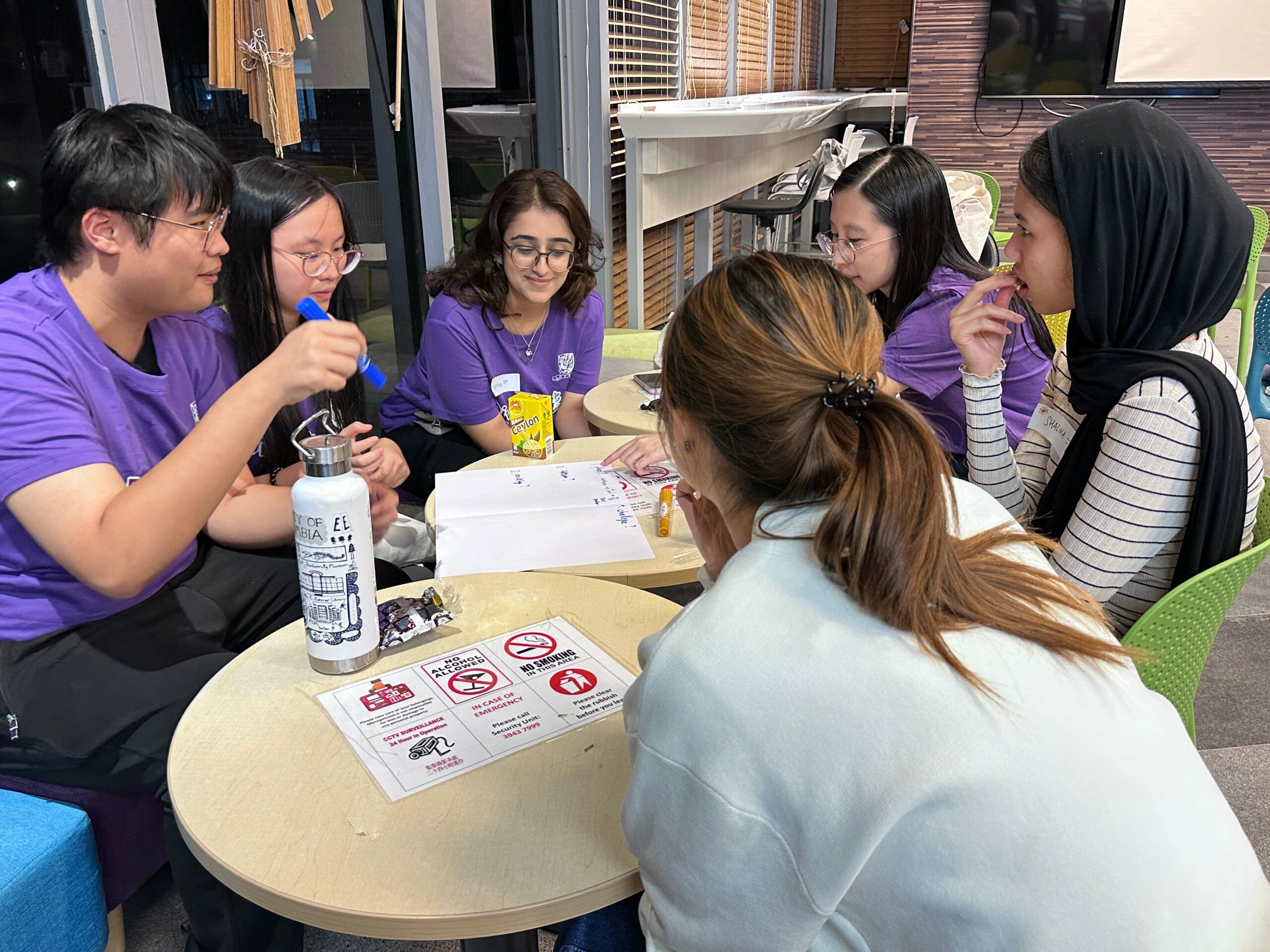 A group of D&I ambassadors sits around a table during a workshop. They are actively engaged in a discussion with papers, markers, and snacks on the table.