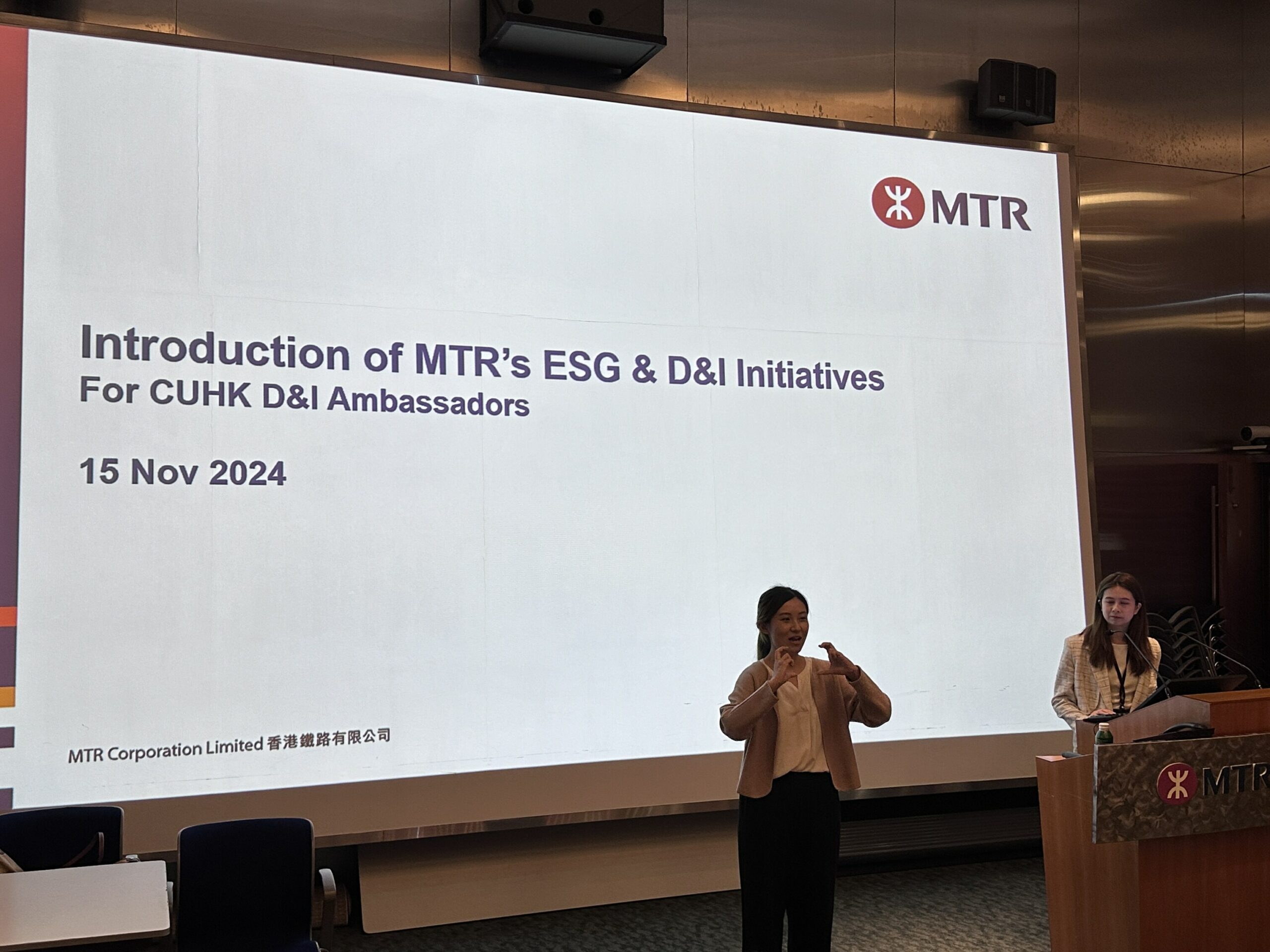 The speaker from MTR are demonstrating the sign language for “Moving train” to the participants, in front of the presentation screen showing the topic “Introduction of MTR's ESG and D&I Initiatives for CUHK Ambassadors.”