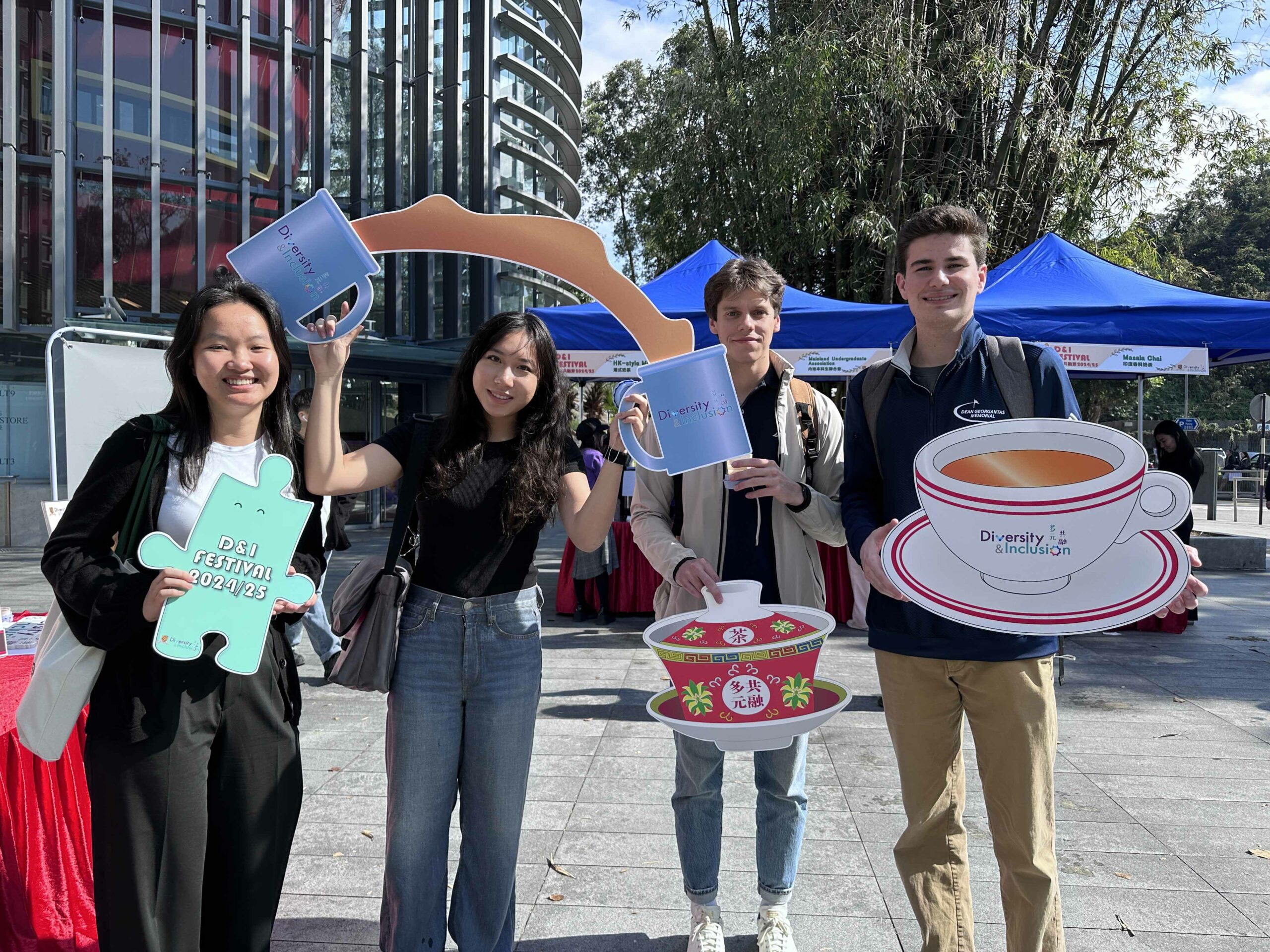 Four students are taking a photo with tea-like props and smiling in front of the event booth.