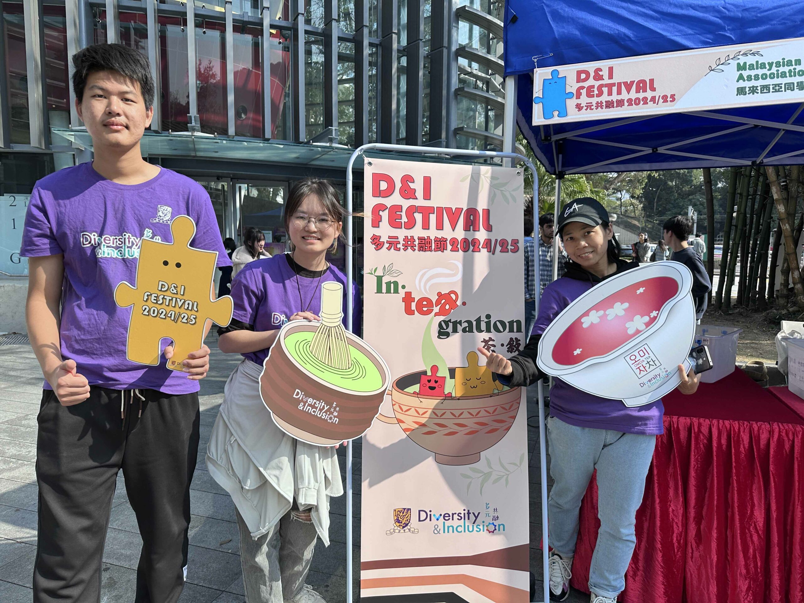 Three D&I ambassadors are standing near the event banner, holding tea-like props and smiling.