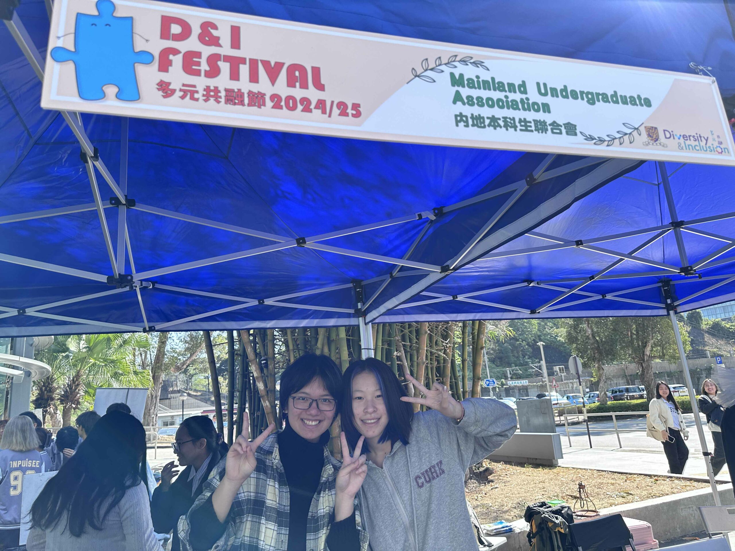 Two students from the Mainland Undergraduates Association are smiling and making “V” signs under a blue tent with a "D&I Festival" banner.