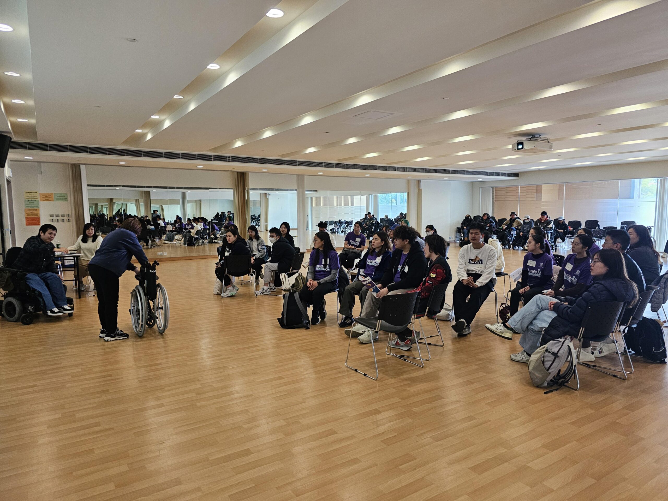 The D&I ambassadors are attentively listening to a tutor who is explaining the proper use of wheelchairs, with some ambassadors trying out the wheelchairs themselves.